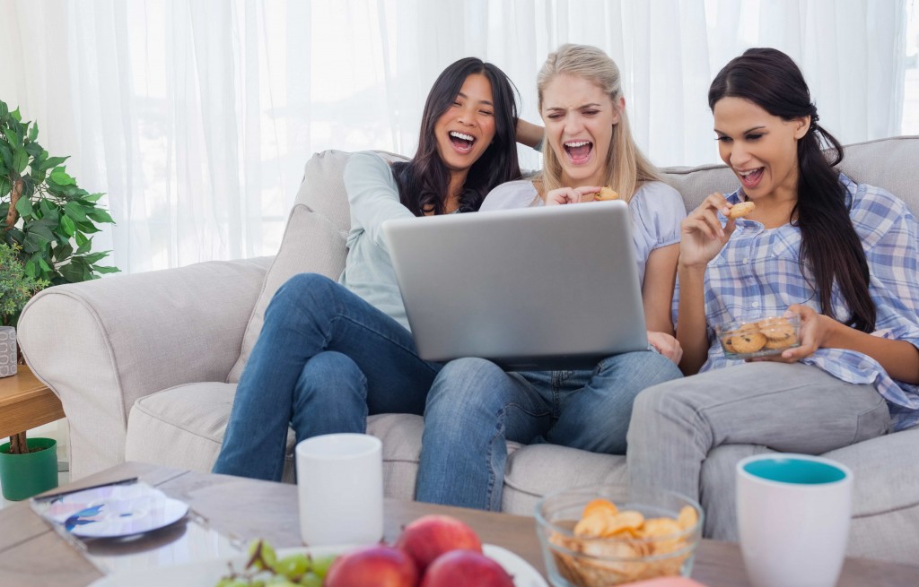Smiling friends looking at laptop together and eating cookies at home on couch